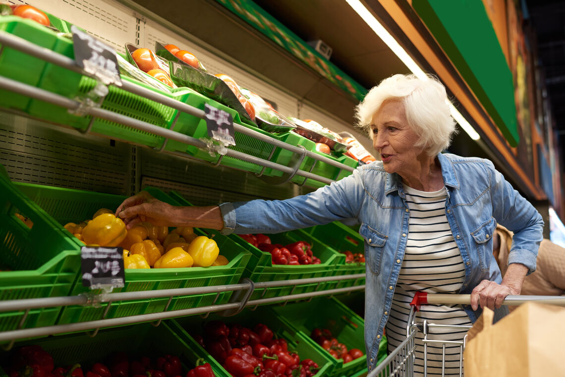 Portrait of modern senior woman with shopping cart  choosing fruits and vegetables in supermarket while enjoying grocery shopping