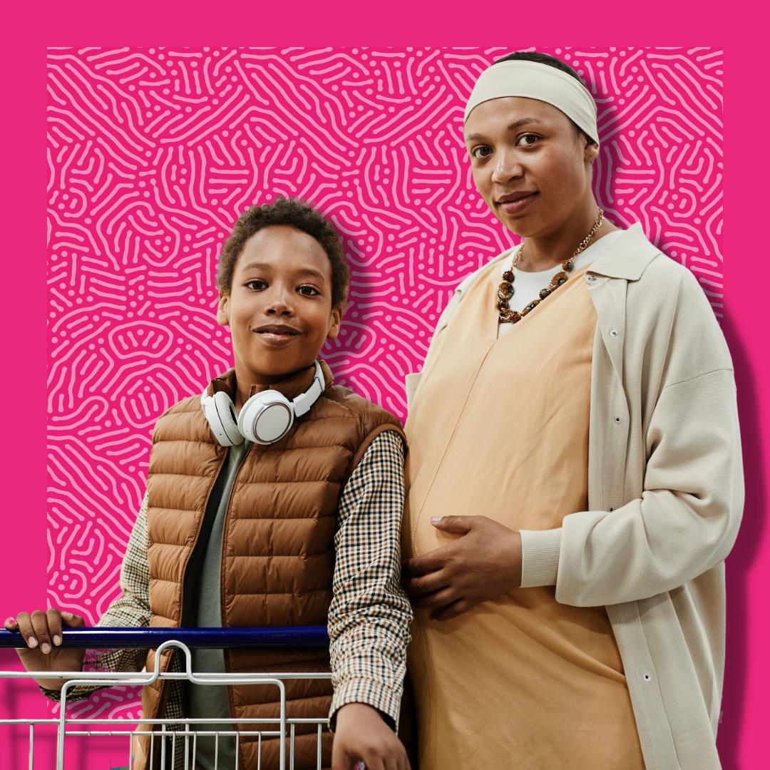 A pregnant mother and her son smile with a shopping cart on a pink patterned background